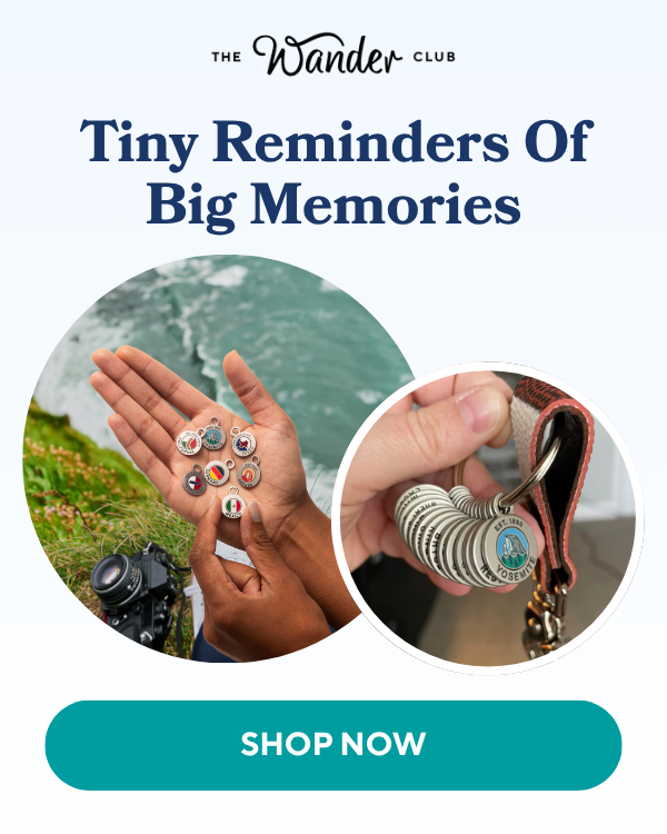 Hands holding various travel charms and a keychain with national park and country flag charms, set against a scenic backdrop.