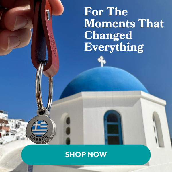 A hand holds a "Greece" keyring with the Greek flag design, set against a blue-domed church in Santorini under a clear sky.
