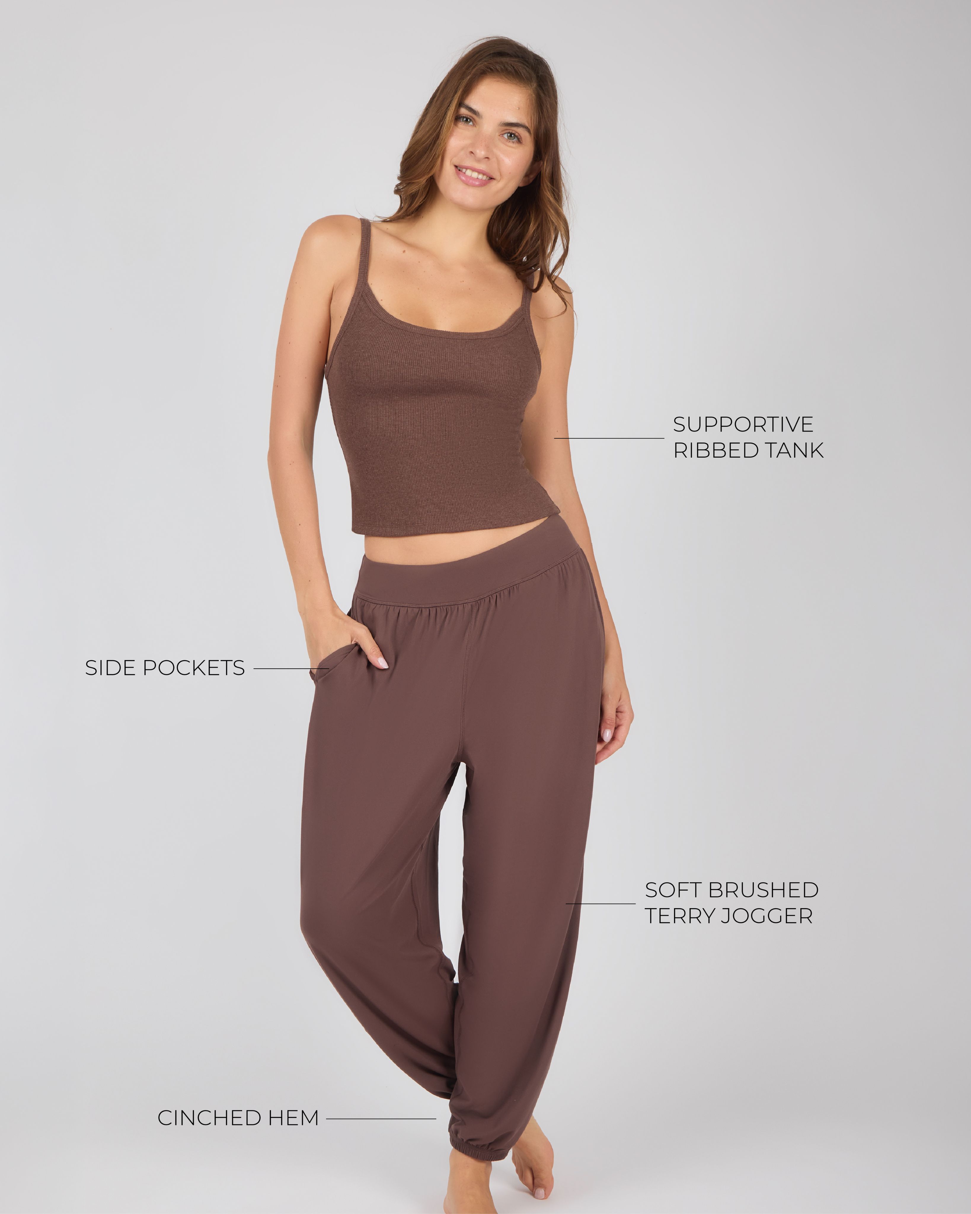 Full-body studio portrait of a woman modeling a brown ribbed tank top and matching brown joggers against a light gray background.