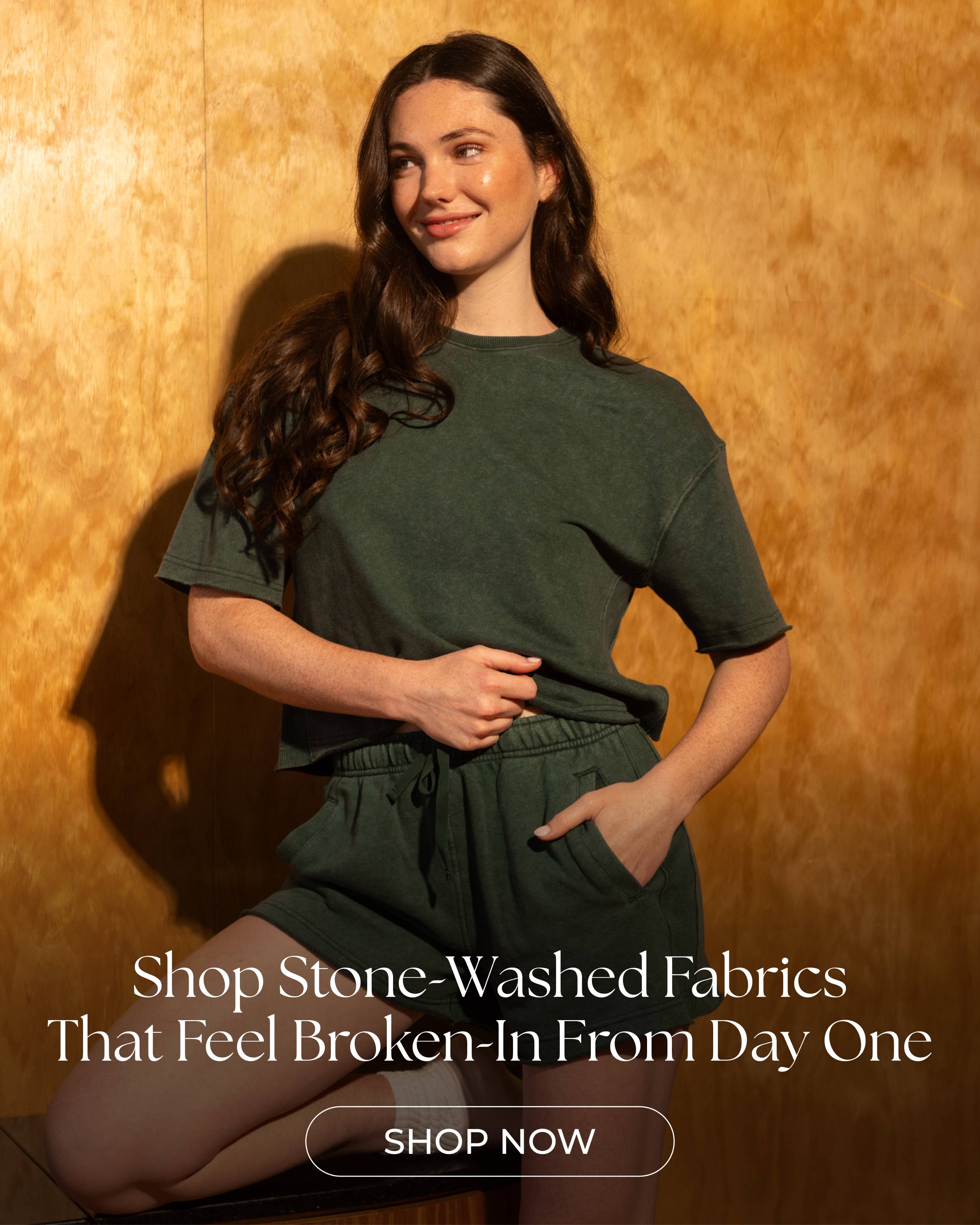 A woman with long brown hair models a dark green stone-washed t-shirt and matching shorts against a wood-paneled wall.