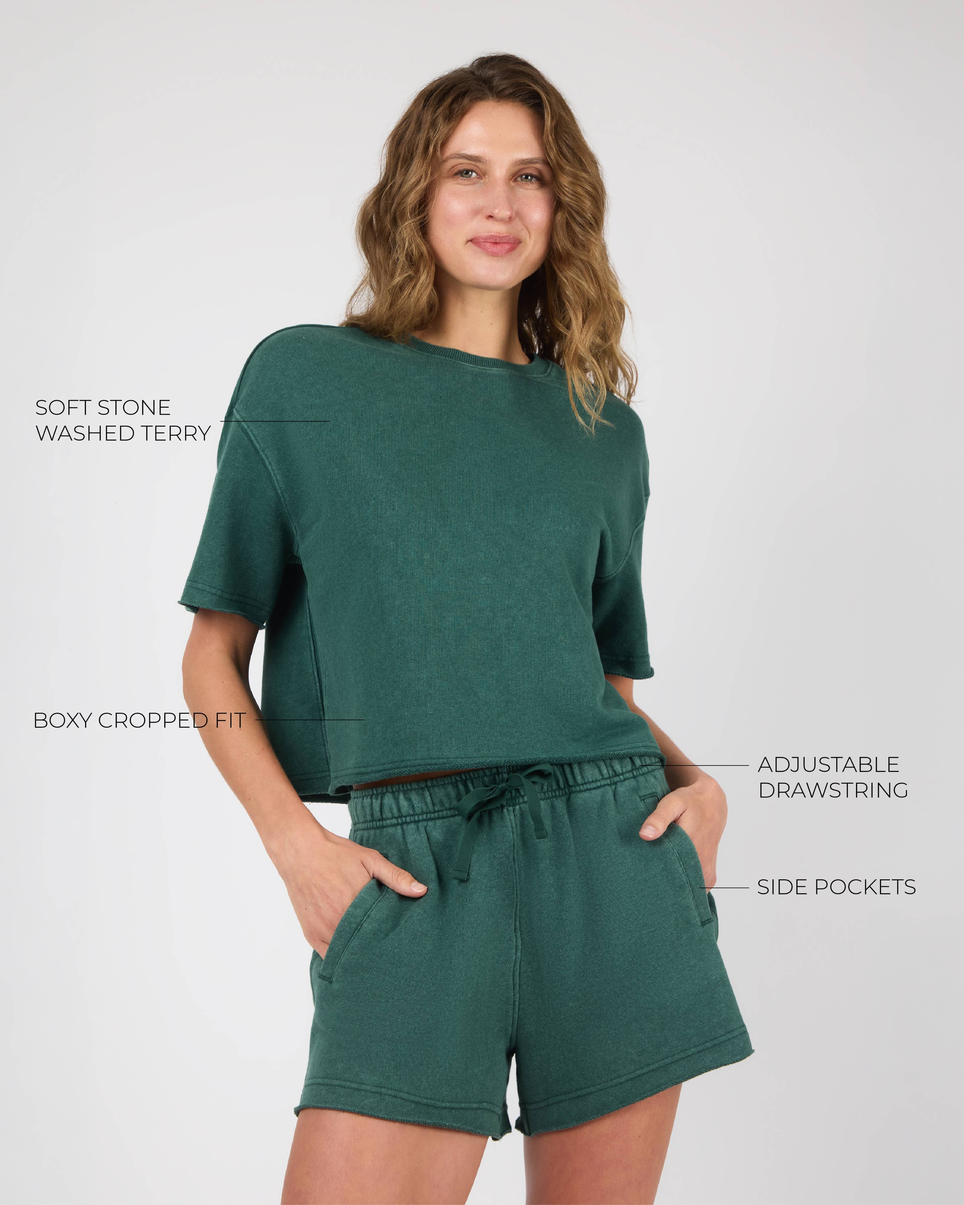 A female model wearing a dark green cropped short-sleeve top and matching shorts against a white background. Text callouts point to product features.